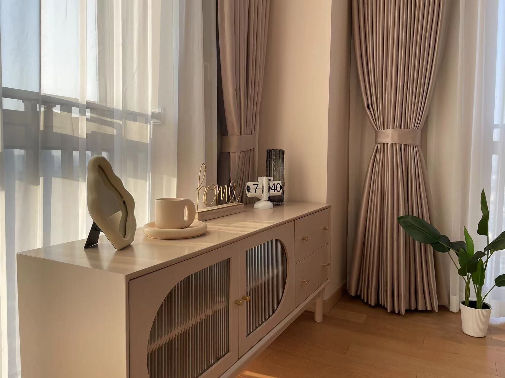 Una sala de estar moderna con una consola beig, una planta en maceta y un ventanal con cortinas translúcidas.
