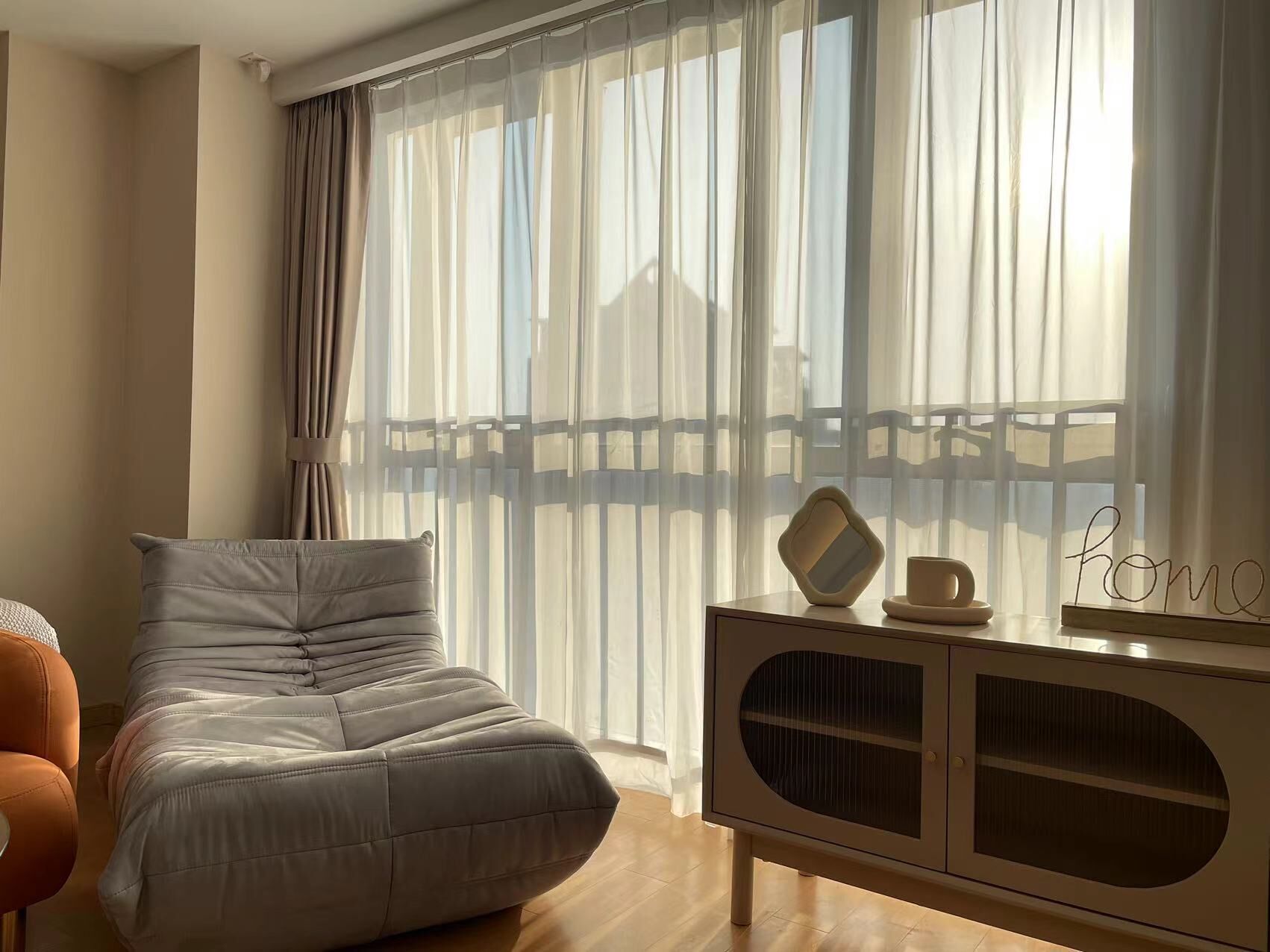 Una sala de estar moderna con un sofá gris, un mueble de madera con puerta de cristal y cortinas ligeras que dejan entrar la luz natural.