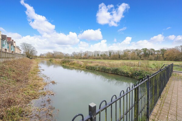 A calm river with a metal railing on the right, surrounded by greenery and trees under a clear sky. - Stylish Flat With Sofa Bed in Malmesbury (Malmesbury)