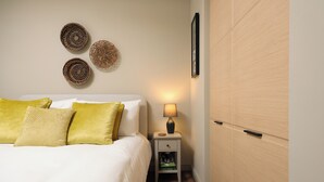 Una cama bien hecha con ropa blanca y almohadas amarillas, una mesita de noche con una lámpara, y una pared decorada con cestas tejidas.