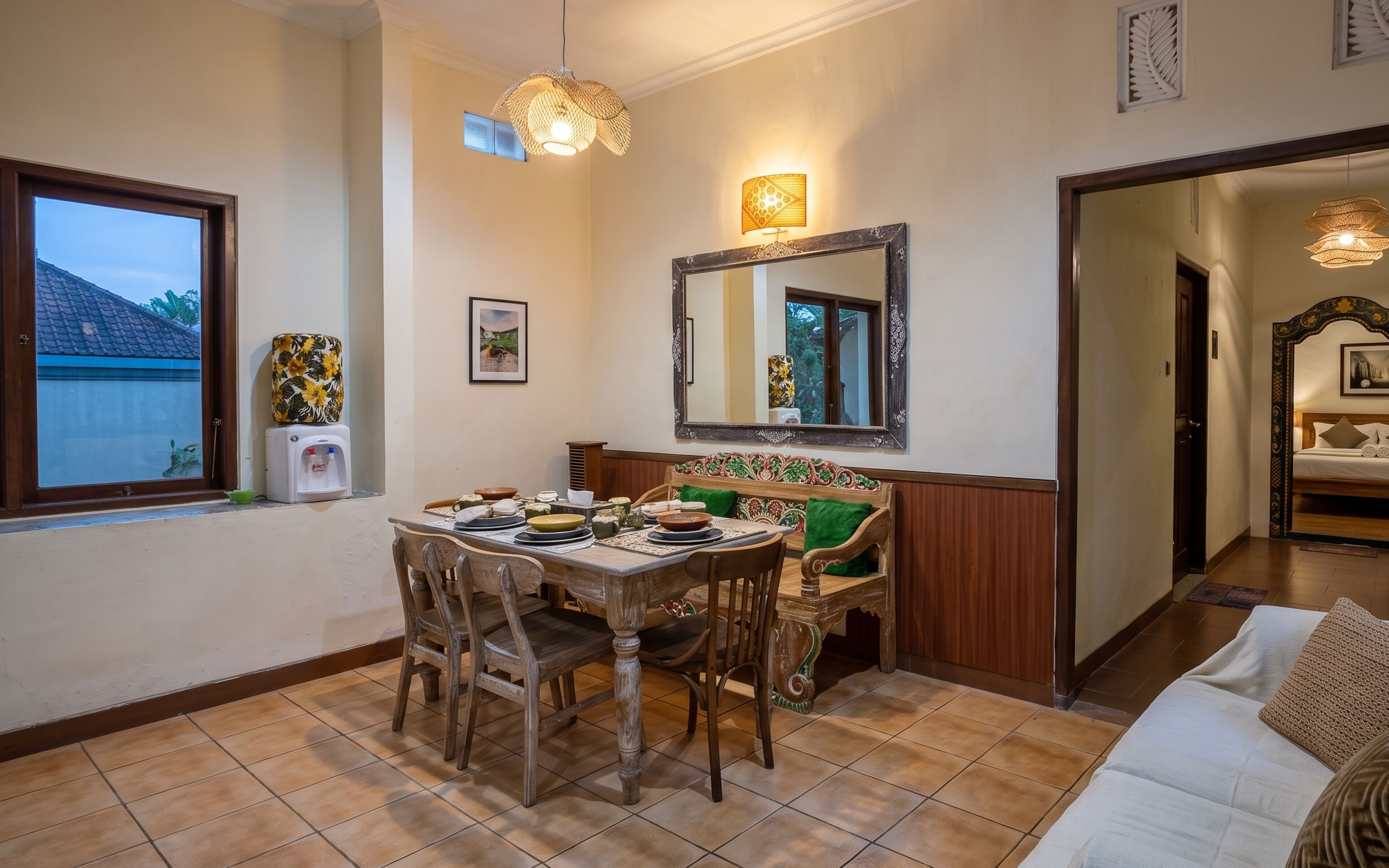 Un coin repas avec une table dressée pour quatre personnes, un canapé, un miroir et une fenêtre donnant sur une maison.