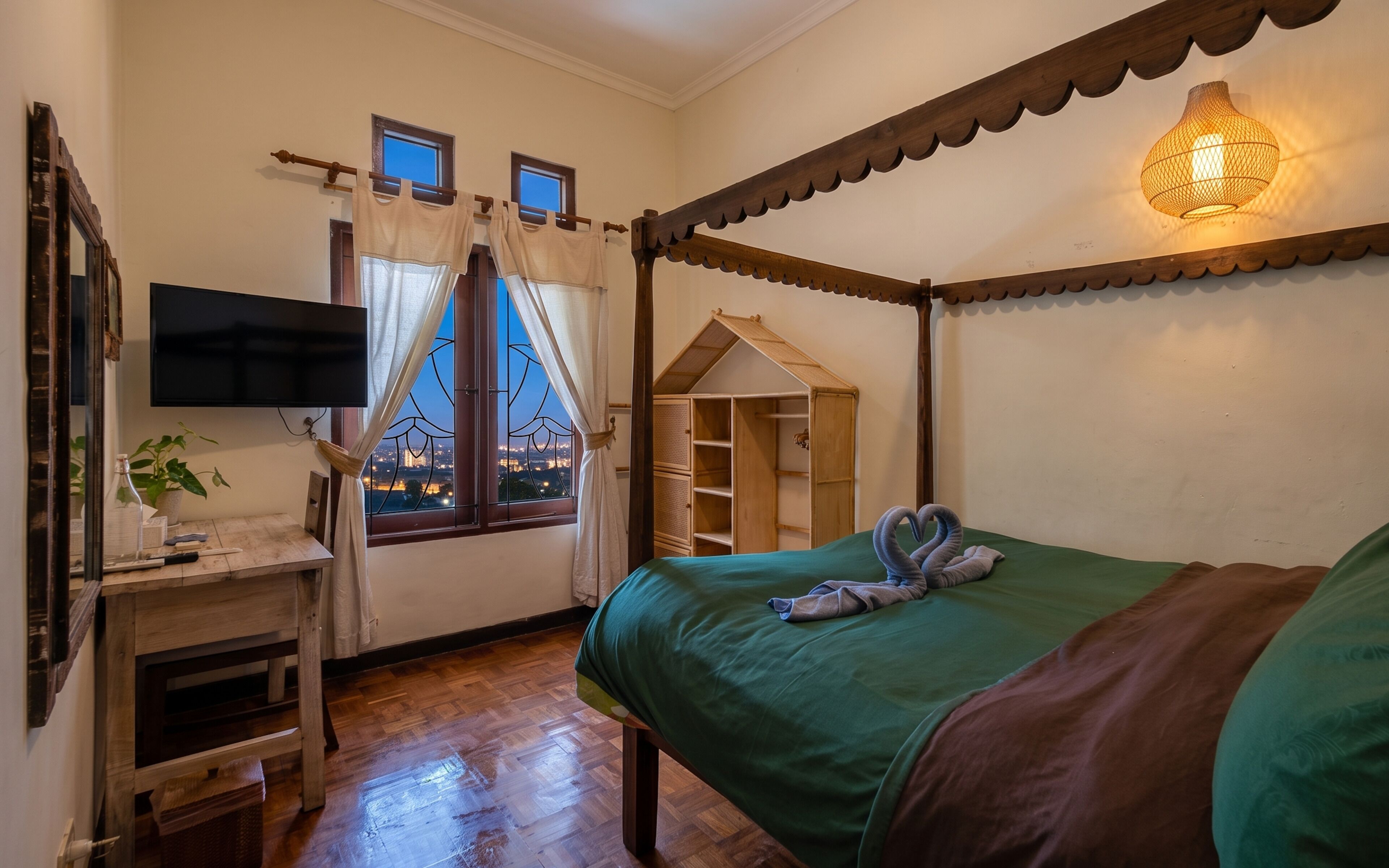 A room with a four-poster bed, a television, a desk with a plant, and a view of the cityscape through the window.