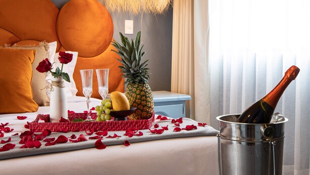 A bed with orange pillows, a vase with a rose, a pineapple, a bottle of champagne, and a fruit bowl.
