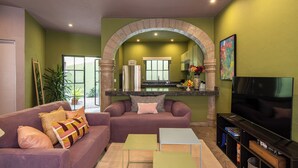 A living room with a sofa, a coffee table, a TV, and a kitchen area visible through an archway.