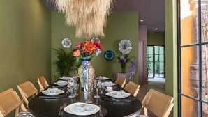 A dining area with a round table set for six, surrounded by wicker chairs. A large vase with colorful flowers is placed at the center of the table.