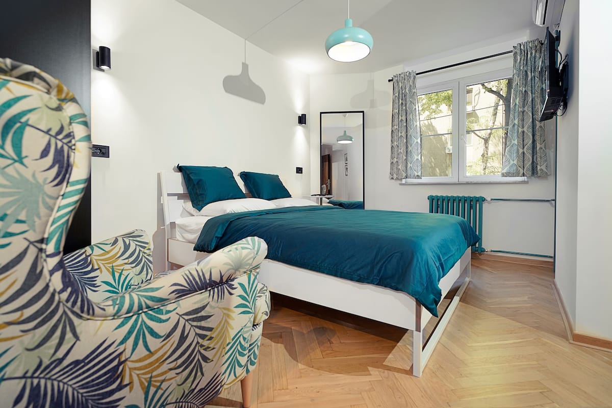 Une chambre moderne avec un lit, une chaise, une fenêtre avec des rideaux, un radiateur et un luminaire au plafond.