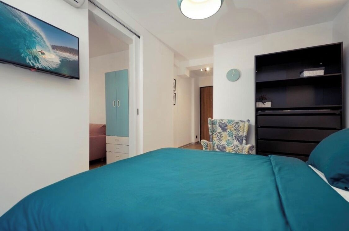 Une chambre avec une couvre-lit turquoise, un téléviseur fixé au mur, une chaise ornée d’un motif de feuilles et une bibliothèque sombre.