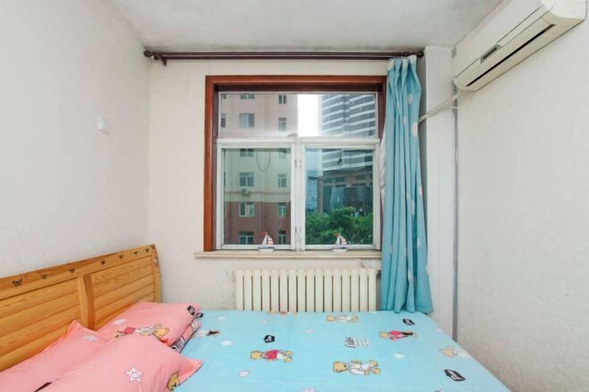 Een slaapkamer met een houten hoofdeinde, een bed met een blauwe sprei waarop tekenfilmfiguren staan afgebeeld, een raam met uitzicht op gebouwen en een airconditioning.