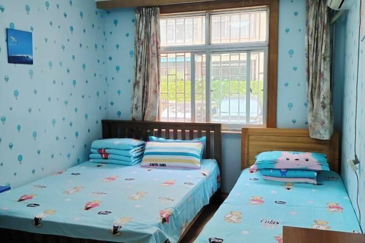 Een slaapkamer met twee bedden, elk met een blauwe dekbedovertrek met tekenfilmfiguren. Er is een raam met gordijnen, en de muren zijn versierd met een patroon van blauwe ballonnen.