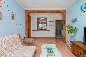 A living room with a sofa, coffee table, and a TV. There is a kitchen area with a window and a plant.
