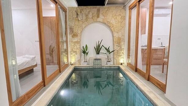 Ein rechteckiger, überdachter Pool mit klarem Wasser, umgeben von Wänden mit Holzzargen und steinernen Akzenten.