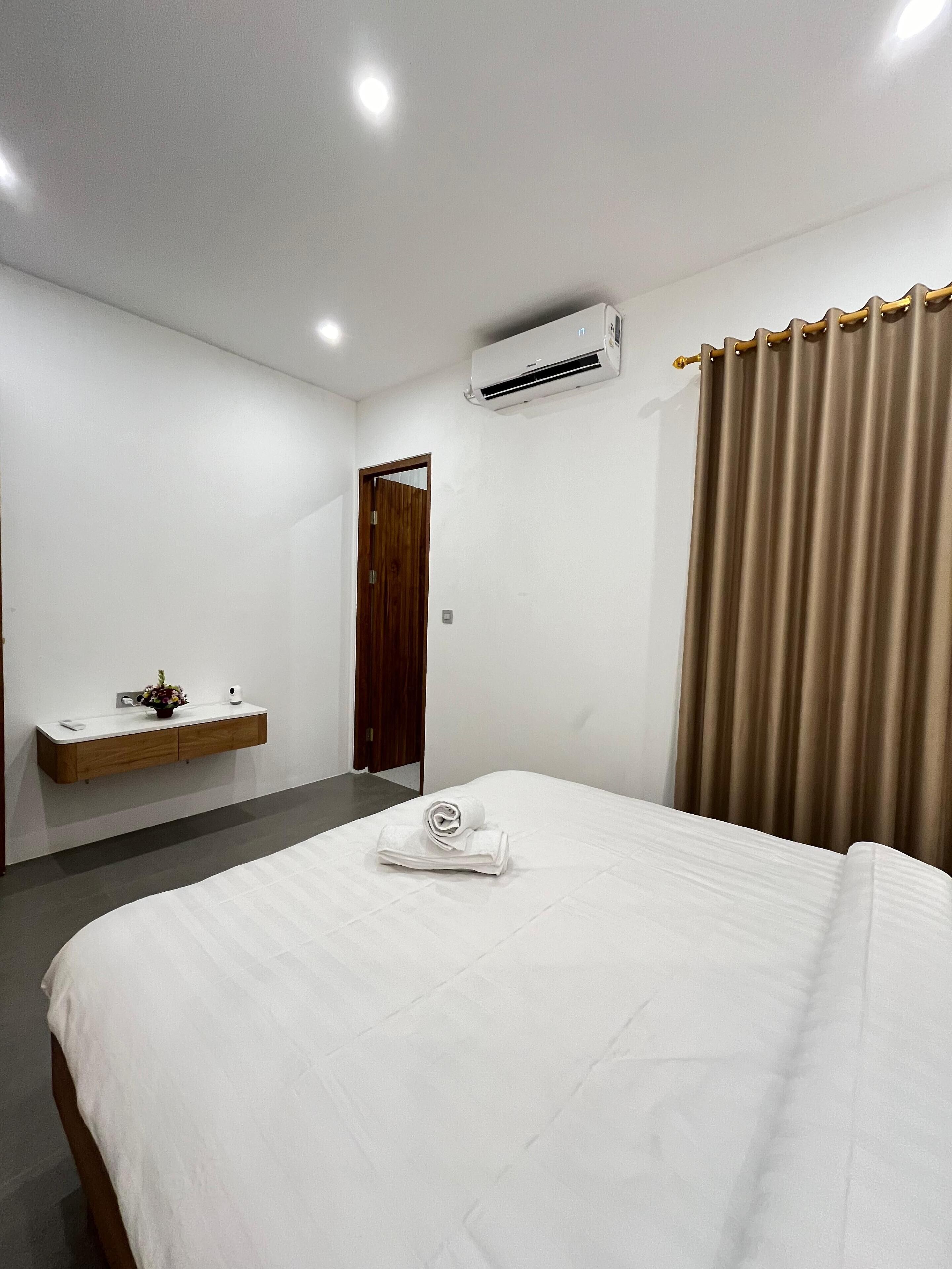 Habitación de hotel con cama, mesita de noche con un jarrón de flores, puerta de madera, unidad de aire acondicionado y cortina.