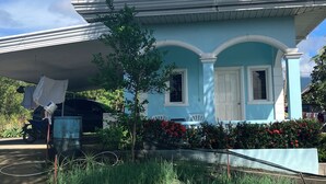 Une maison bleu clair, avec une porte et des fenêtres blanches, entourée d’un jardin où poussent différentes plantes et fleurs.
