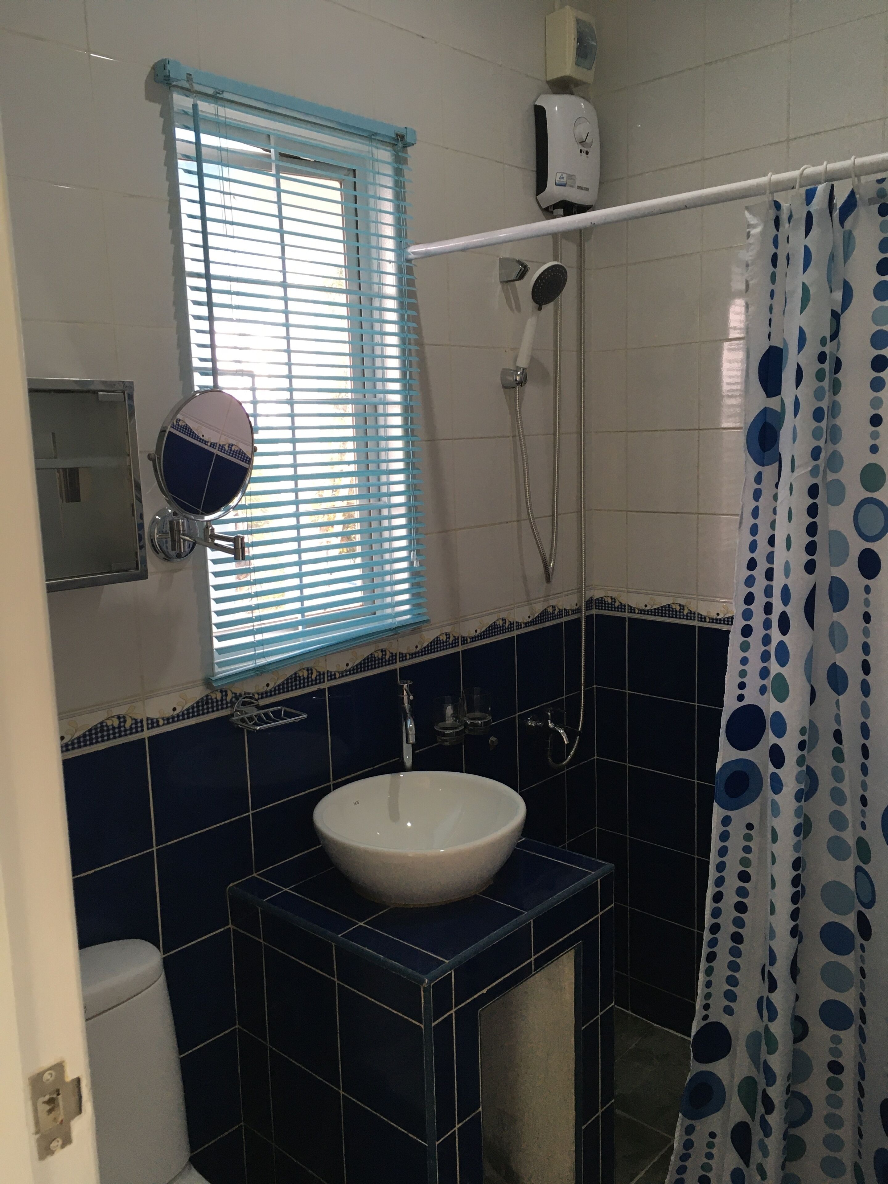 Un bagno con lavandino bianco, piastrelle blu, una finestra con persiane, una doccia e una tenda a pois.