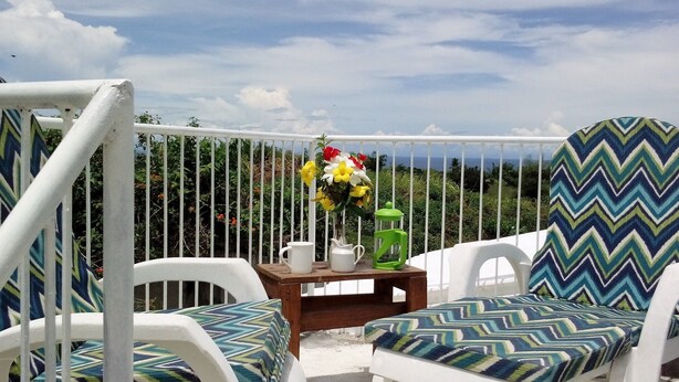 Un balcon avec des chaises blanches et une table, un vase de fleurs et une vue sur un paysage luxuriant.