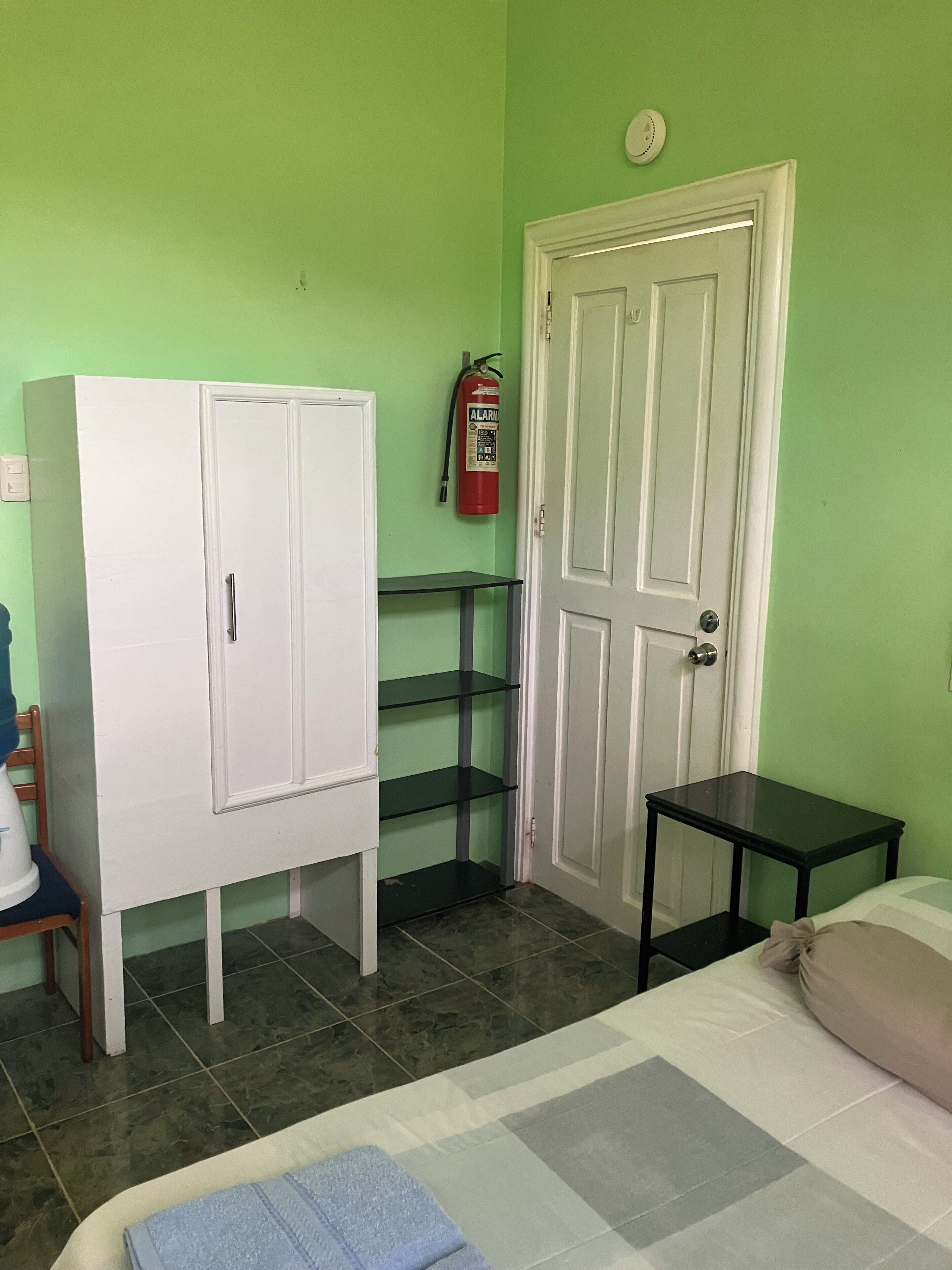Una habitación con una cama, un armario blanco, una mesita negra pequeña, un extintor de incendios y una pared verde.