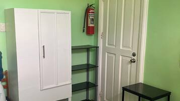 Una habitación con una cama, un armario blanco, una mesita negra pequeña, un extintor de incendios y una pared verde.