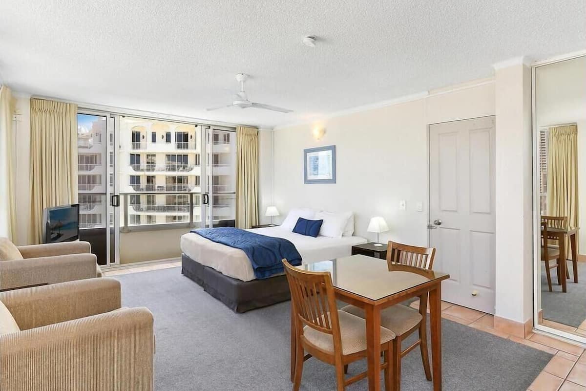 A hotel room with a bed, a desk, and a chair. There is a window with curtains, a door, and a ceiling fan.