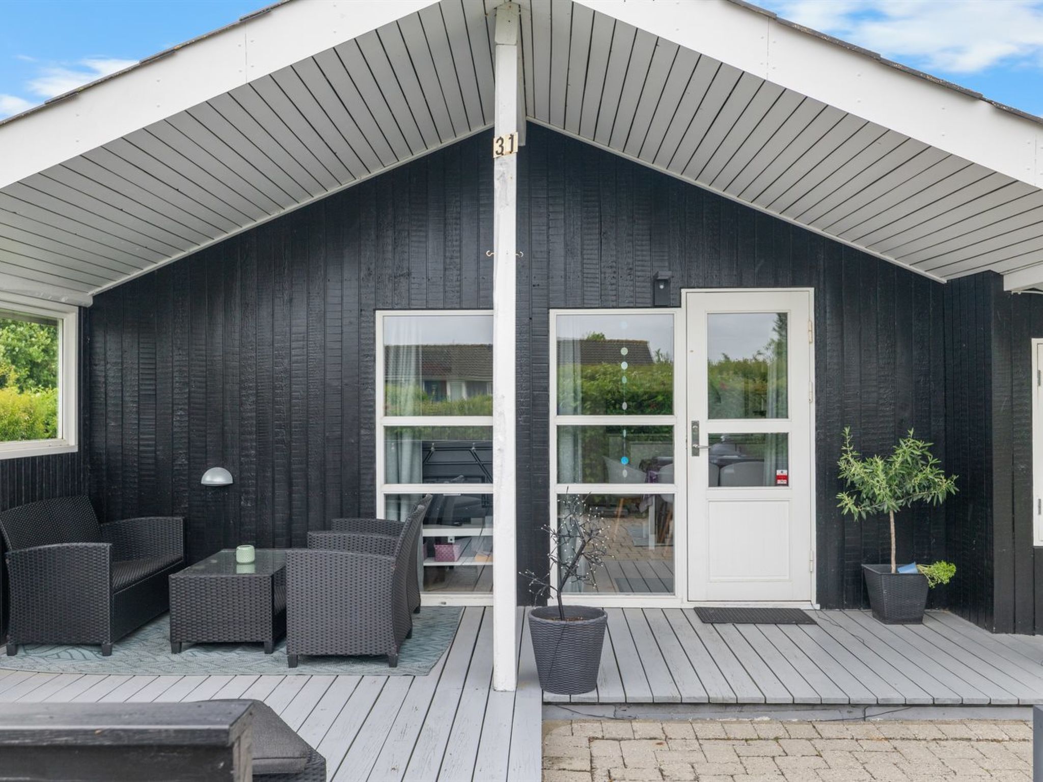 A covered outdoor area with a dark wooden exterior, a white door, and a seating area with wicker furniture.