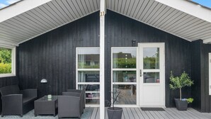 Un espace extérieur abrité, doté d’une façade en bois sombre, d’une porte blanche et d’un coin salon avec des meubles en osier.