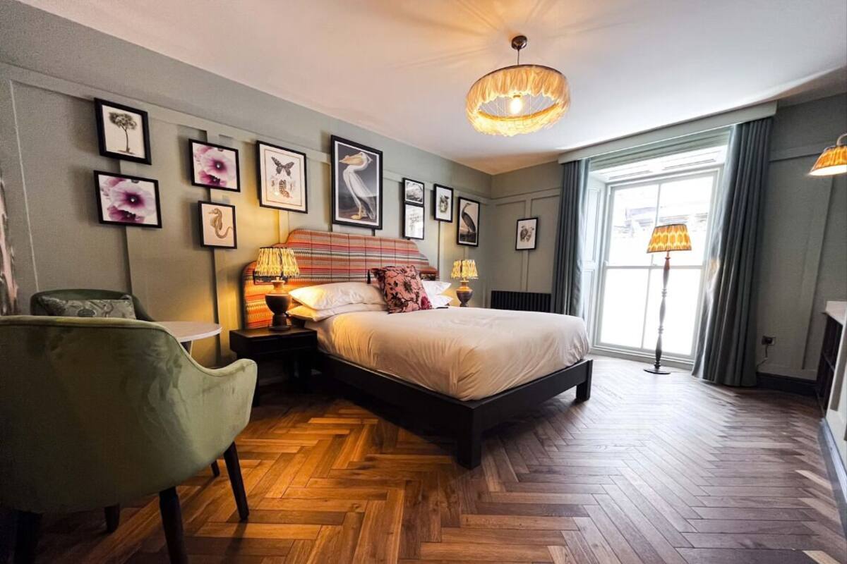 A bedroom with a large bed, a green armchair, a wooden herringbone floor, and framed artwork on the walls.