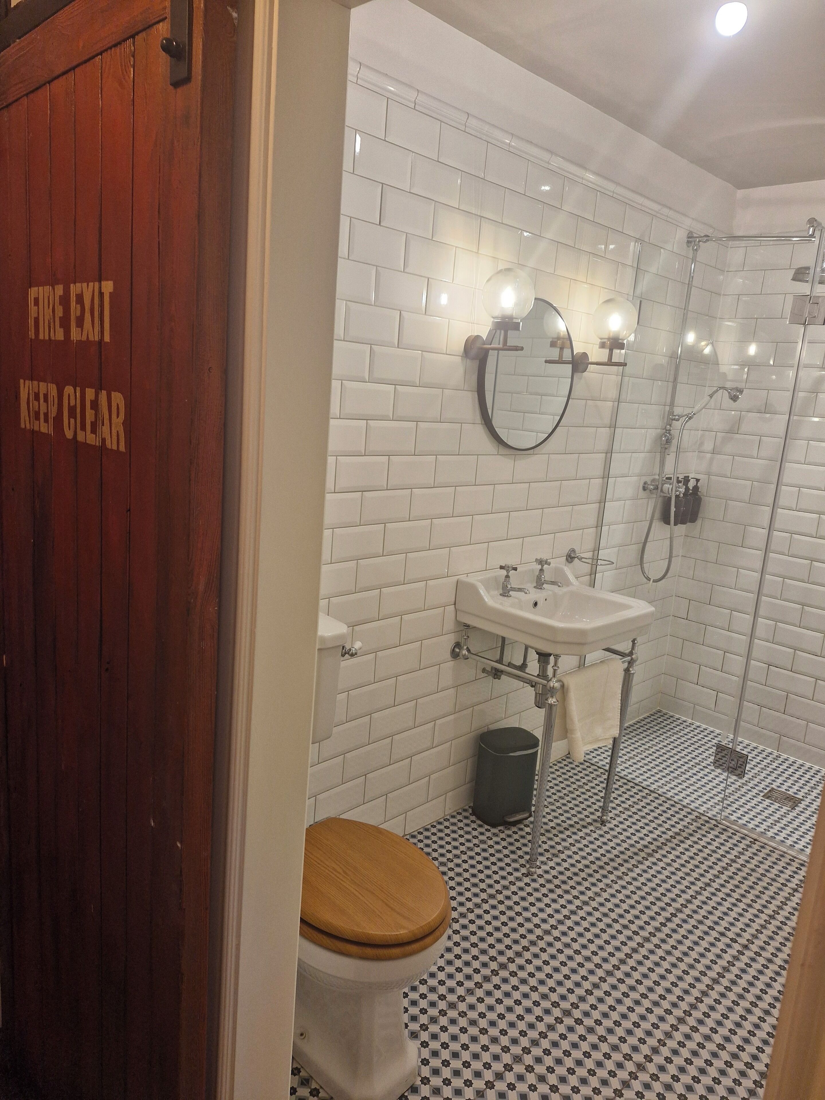 Un bagno con piastrelle bianche tipo metro, una porta in legno con segnale di uscita di sicurezza, un water, un lavandino, uno specchio e una doccia.