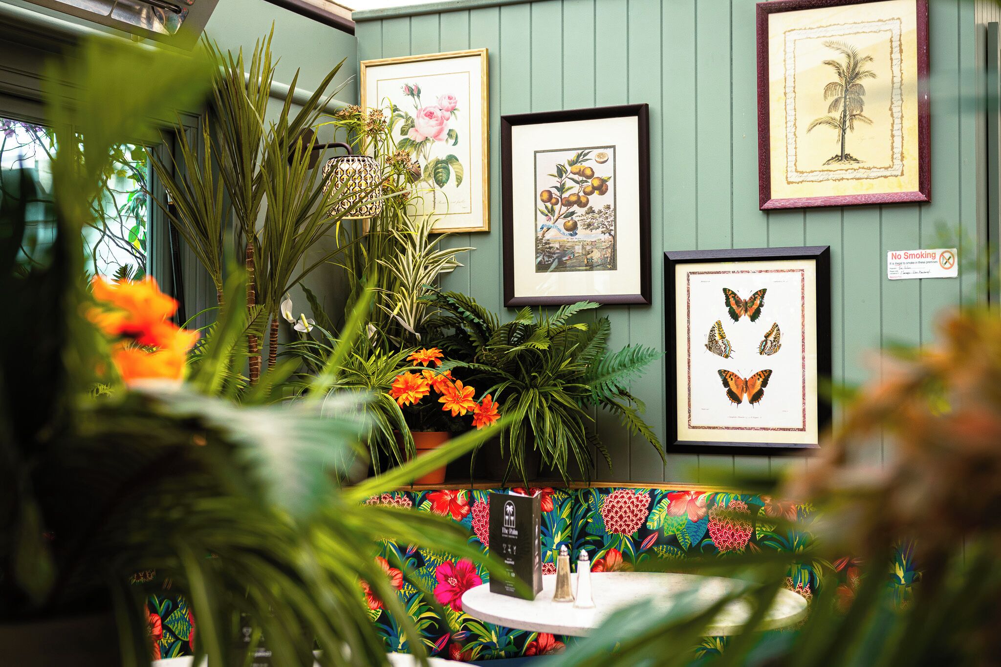 A room with framed botanical prints on the wall, a round table with salt and pepper shakers, and a colorful floral-patterned bench.