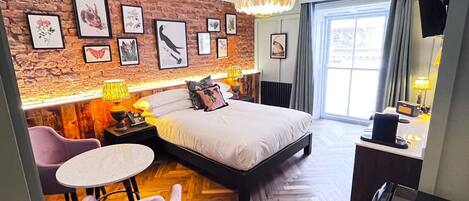 Una habitación de hotel moderna con una pared de ladrillo, una cama con duvet blanco, un escritorio con lámpara, una silla y un ventanal grande.
