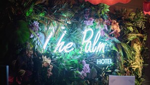Un mostrador de recepción con un letrero de neón que dice "The Palm Hotel" en medio de abundante follaje y plantas tropicales.