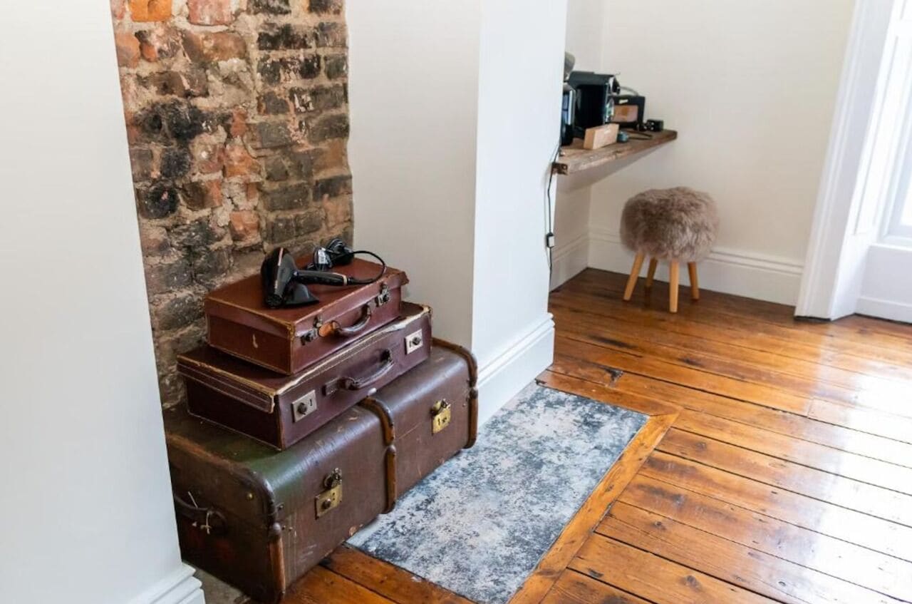 A room with a brick wall, wooden floor, and a small stool. There are suitcases stacked against the wall and a shelf with items on it.