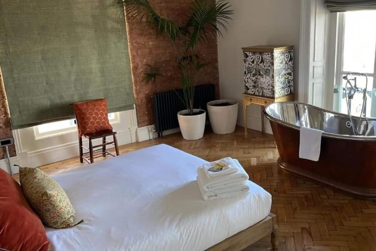 A hotel room with a large bed, a bathtub, a potted plant, and a view of the outdoors.
