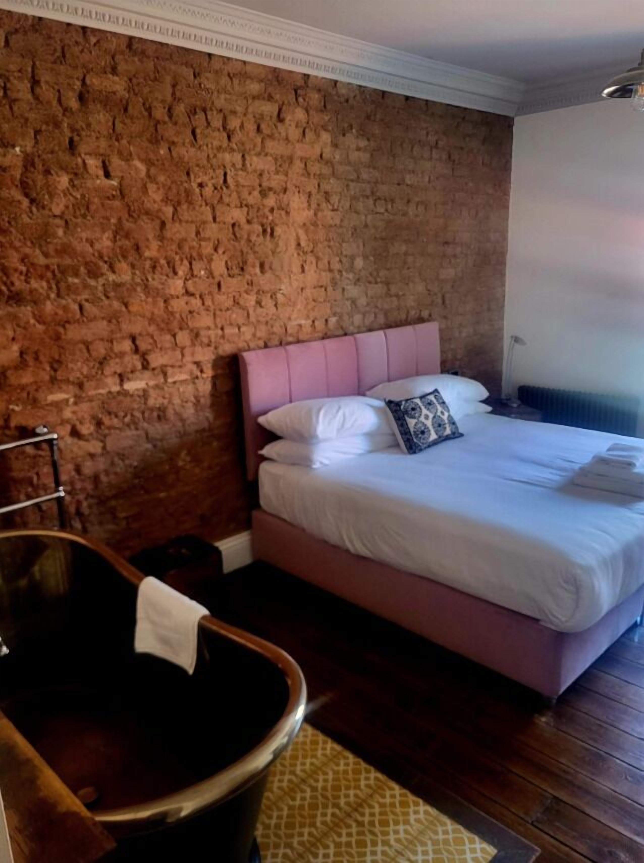 A room with a brick wall, a wooden bathtub, a bed with white bedding, and a small bedside table.