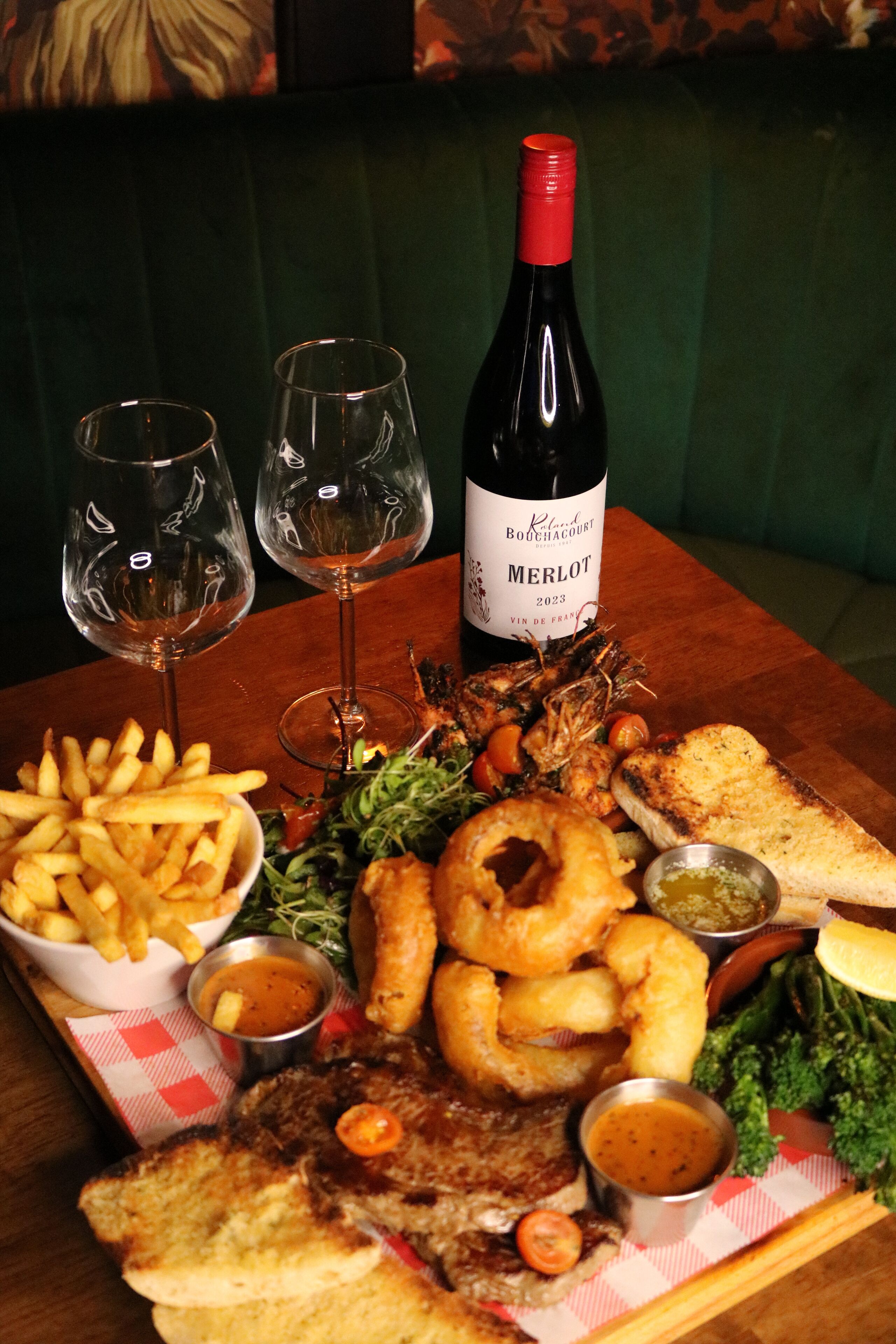 Una mesa de comedor con una botella de Merlot, dos copas de vino vacías, y una variedad de comida que incluye papas fritas, ensalada, aros de cebolla y carne a la parrilla.