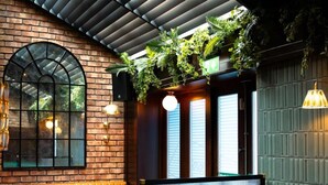 Interior de un restaurante con paredes de ladrillo, mesas de madera, asientos tapizados en verde y plantas colgantes.