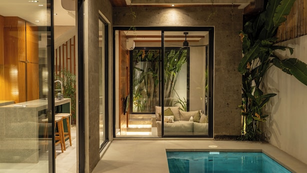 A modern indoor pool area with a glass wall, a wooden bench, and a sofa in the background. - Nanna Villa Unit 4 by Betterplace (Canggu)