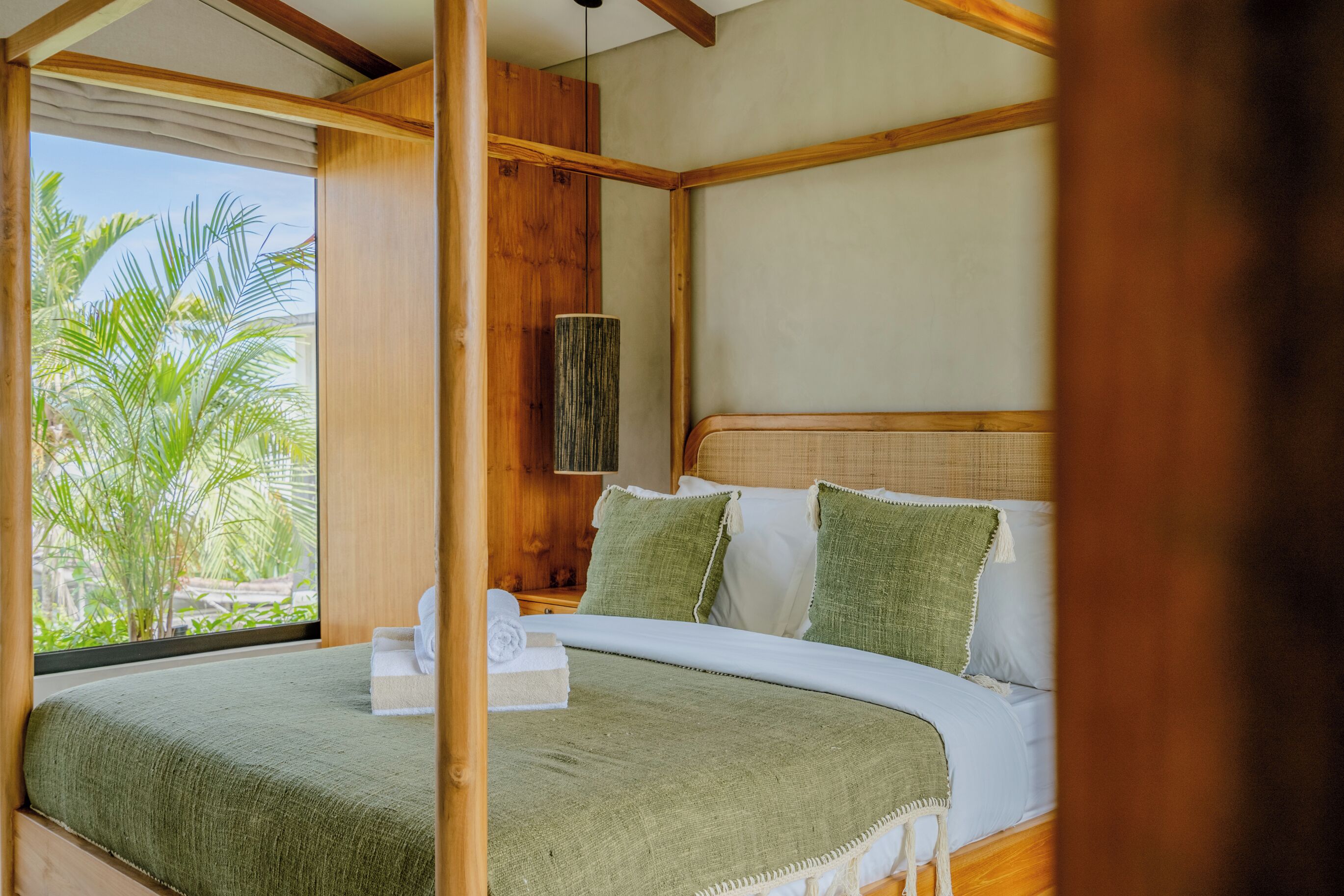 A room with a bed, a window with a view of palm trees, and a wooden ceiling.