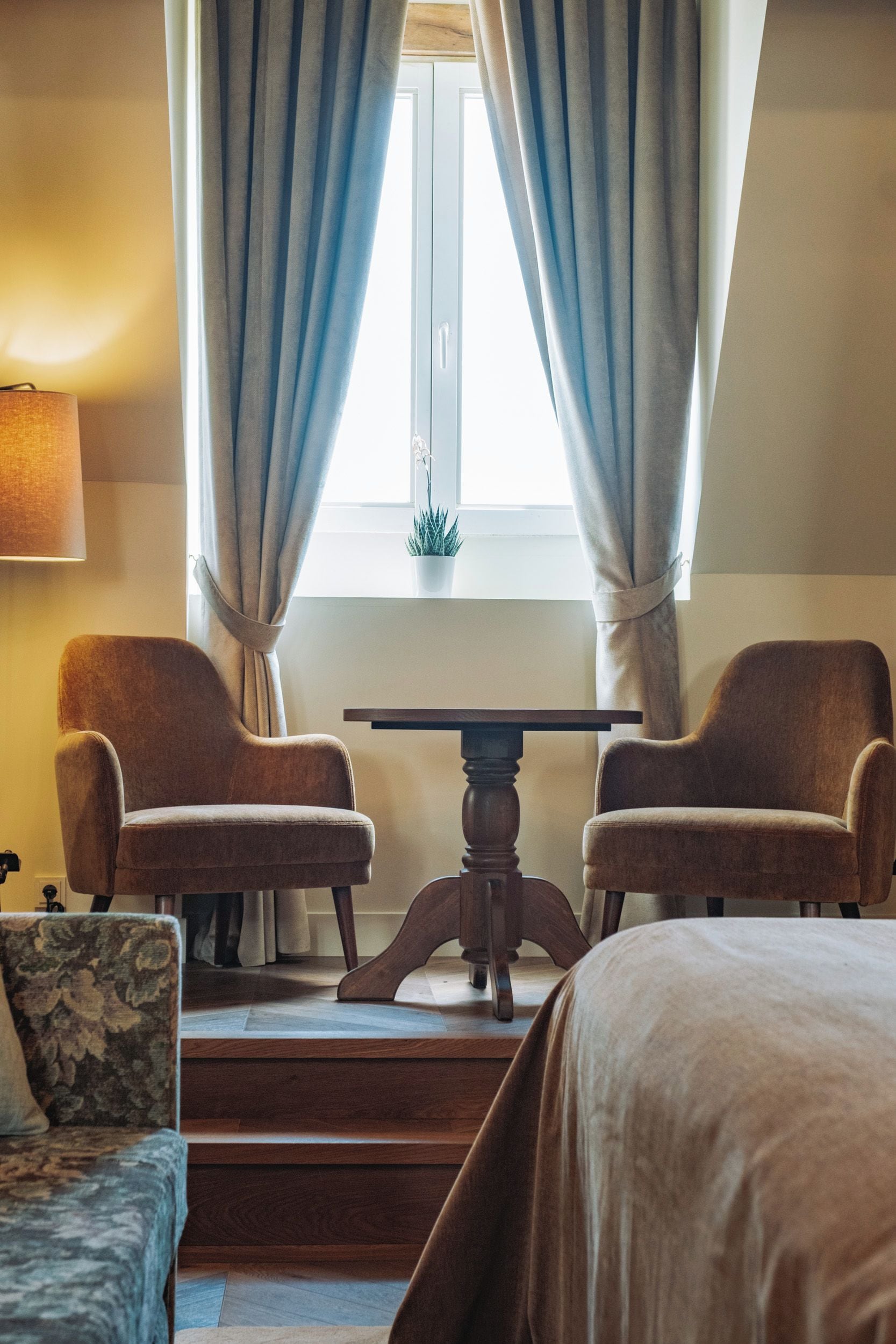Et hotelværelse med to lænestole, et lille bord, en seng og et vindue med gardiner.