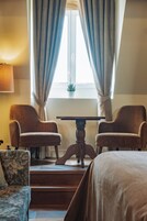 Quarto de hotel com duas poltronas, uma mesa pequena, uma cama e uma janela com cortinas.