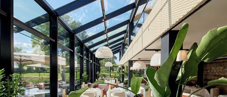 A restaurant with a glass ceiling, outdoor seating, and large green plants.