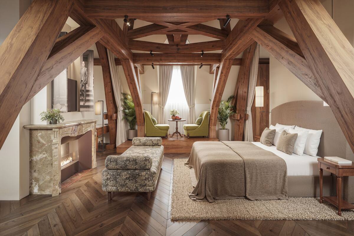 Une chambre spacieuse située dans les combles, avec des poutres apparentes, un grand lit, deux fauteuils, une petite table et une cheminée.