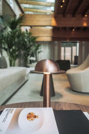 Una lámpara moderna con base de cobre y pantalla blanca, sobre una mesa en un espacio interior bien iluminado, con techo de madera y zona de estar al fondo.