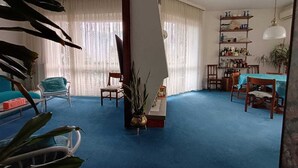 Uma sala de estar com carpete azul, uma área de jantar com mesa e cadeiras, e uma estante cheia de garrafas.