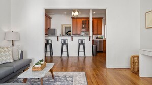 A modern living room with a sofa, coffee table, and bar stools leading to a kitchen with wooden cabinets and a central island.