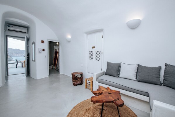 Uma sala de estar moderna com um sofá branco, uma mesa de centro de madeira e vista para o mar através de uma porta aberta.