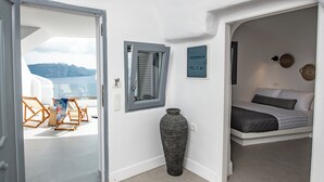 A room with a bed, a large vase, and a view of the sea through a window. - Grand Cave Villa with Outdoor Hot Tub (Thira - Santorini)