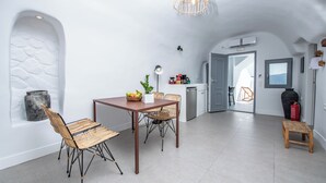 A modern kitchen with a dining area, featuring a stone wall, a wooden table, wicker chairs, and a hanging light fixture. - Grand Cave Villa with Outdoor Hot Tub (Thira - Santorini)