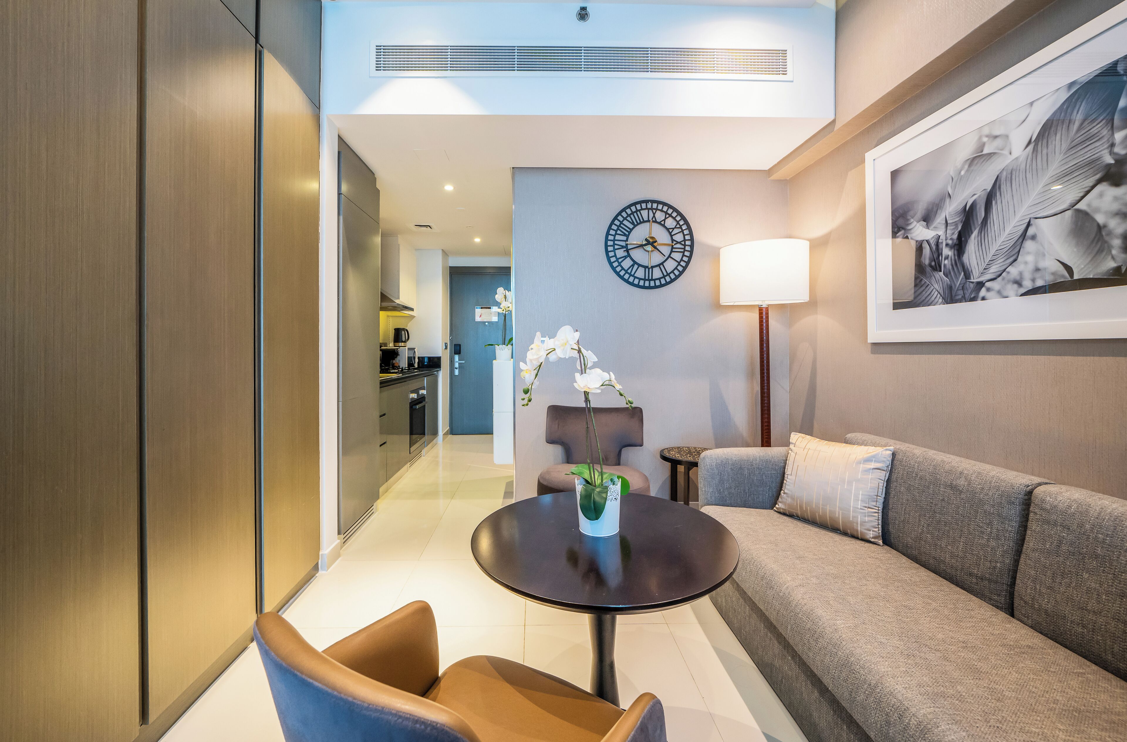 Une chambre d’hôtel moderne, avec un canapé, une table ronde ornée d’un vase de fleurs, une horloge au mur et un tableau encadré.