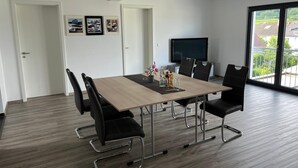 Un comedor moderno con una mesa de madera y sillas negras, un televisor de pantalla plana y un balcón con vistas a casas.