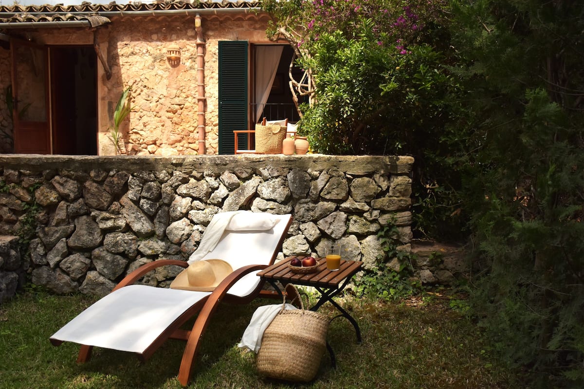 A stone-walled outdoor seating area with two lounge chairs, a small table with a basket of fruit and a glass of orange juice, and a woven basket.