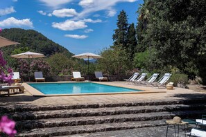 Area piscina con lettini e ombrelloni, circondata da alberi e una montagna sullo sfondo.